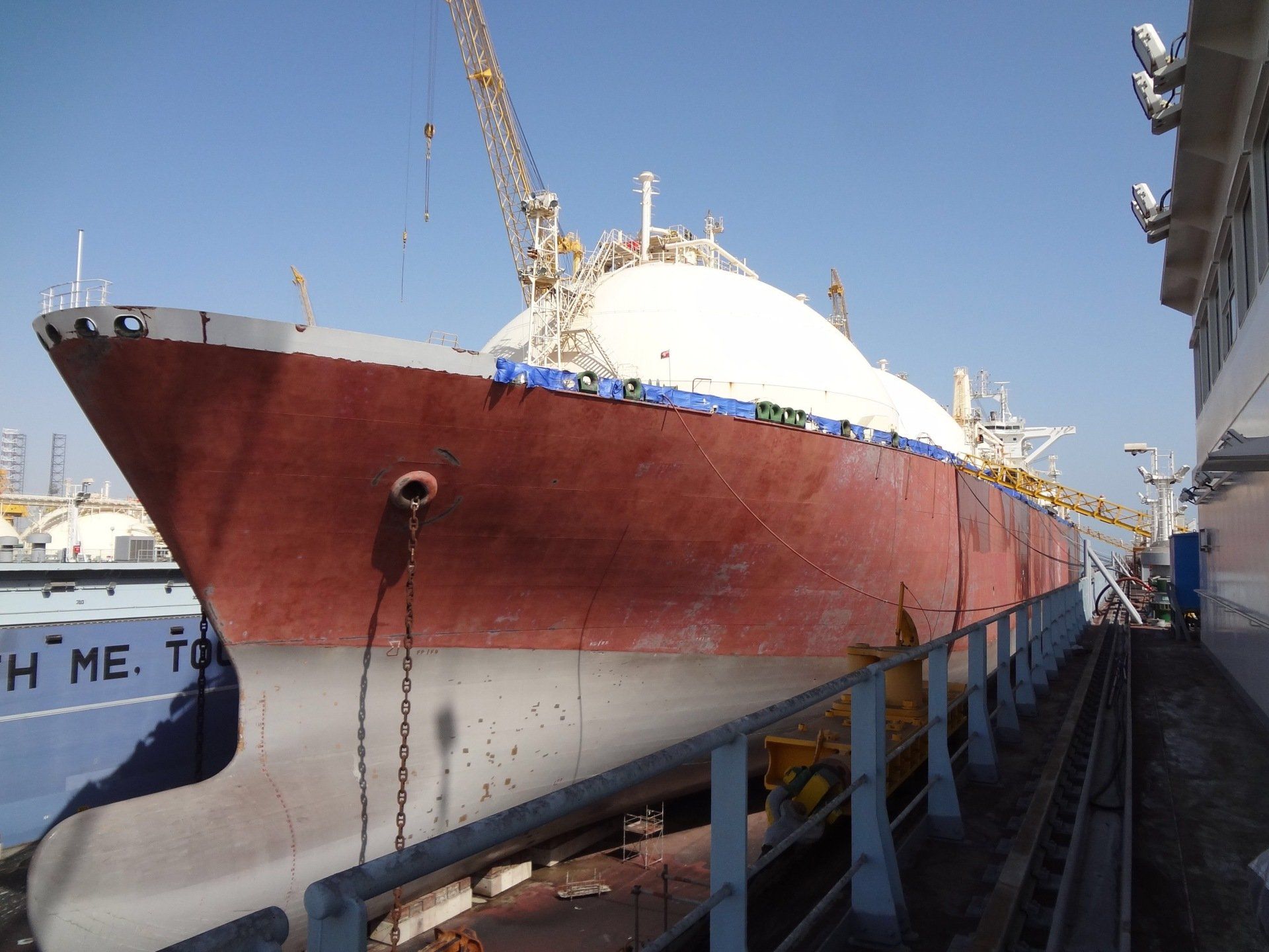 LNG tanker being repaired in dry dock, with a crane overhead and blue sky.