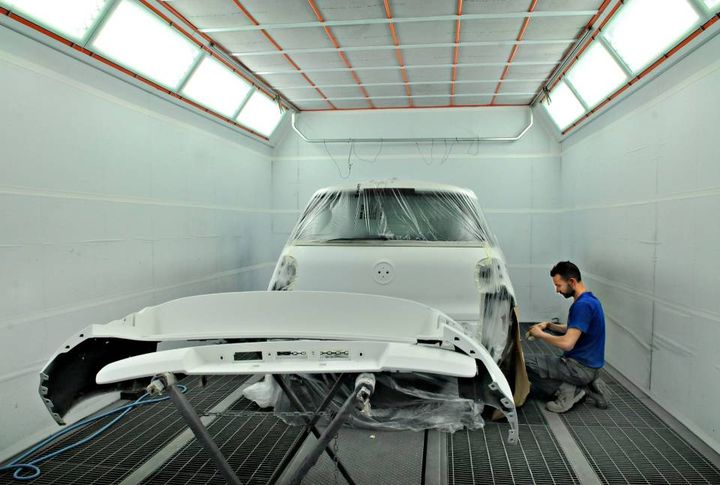 Una persona carteggia la carrozzeria bianca di un'auto in una cabina di verniciatura, con un paraurti staccato in primo piano.