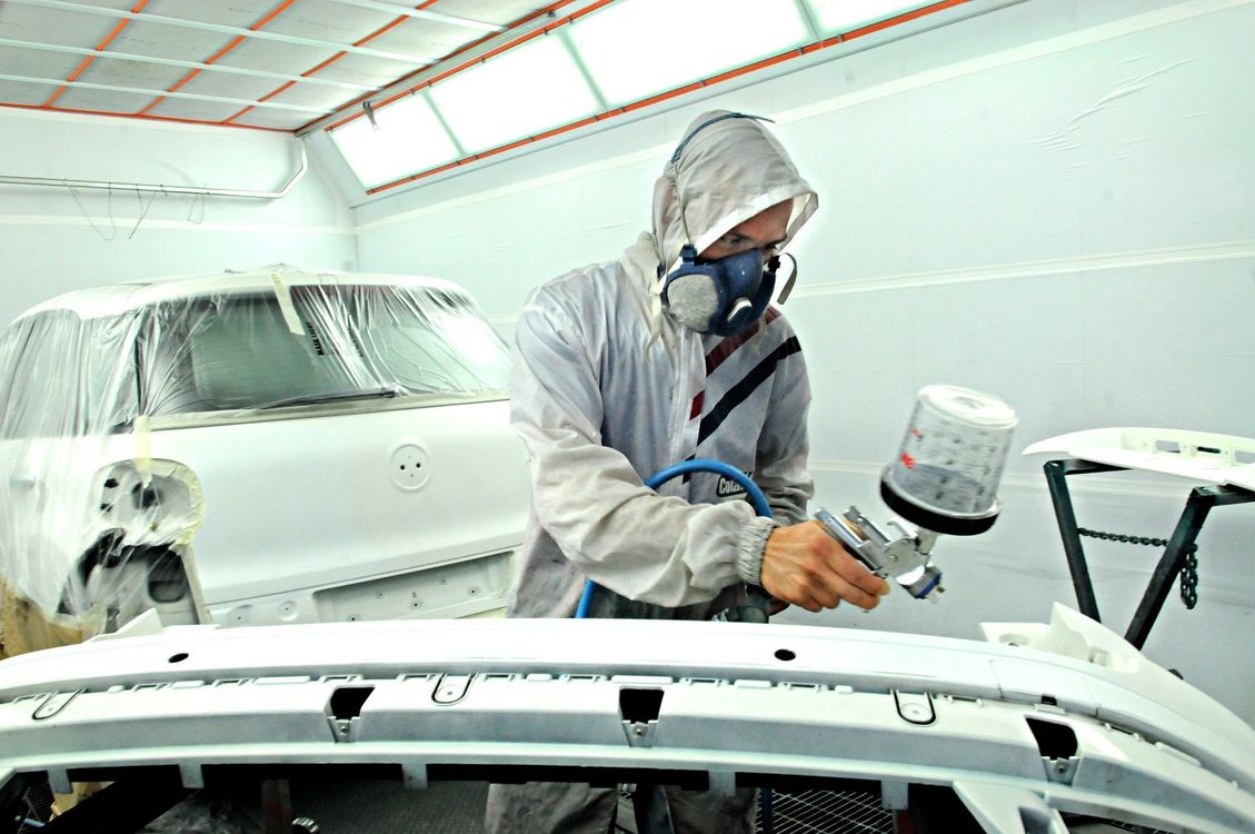 Una persona con la maschera respiratoria vernicia a spruzzo la carrozzeria bianca di un'auto in un'officina.
