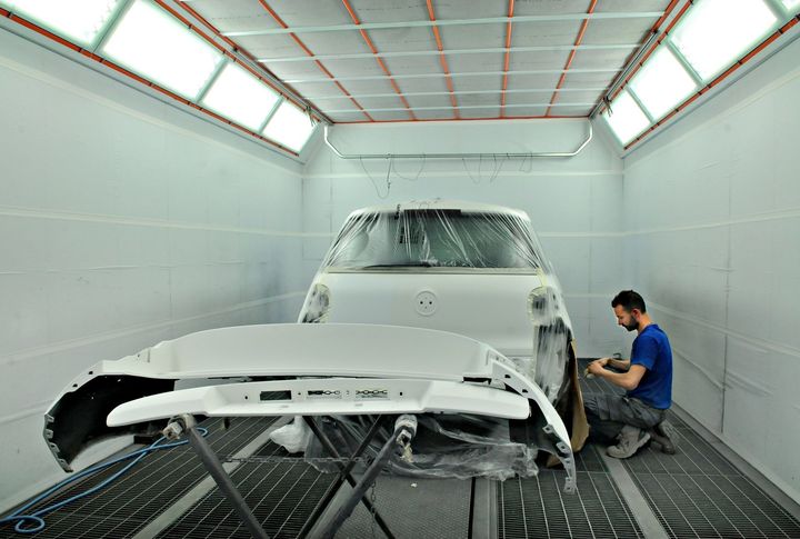 Un carrozziere carteggia un'auto bianca in una cabina di verniciatura con il paraurti anteriore staccato.