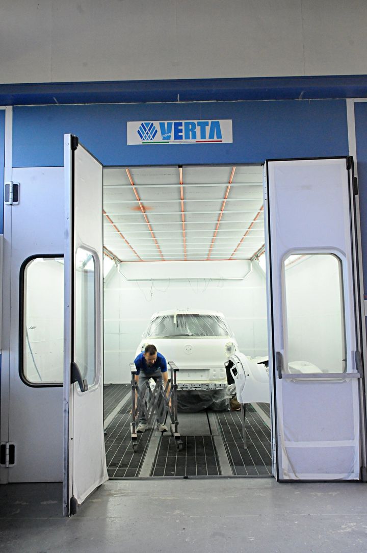 Area di lavaggio auto con un'auto all'interno e un addetto che la sta risciacquando, vista attraverso le porte blu aperte.
