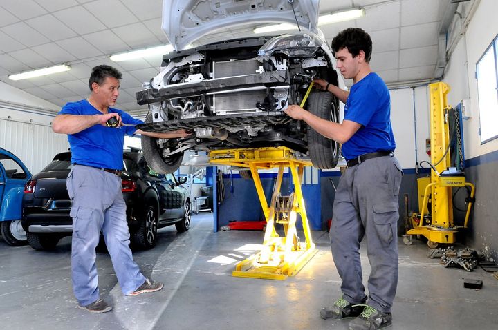 Due meccanici in camicia blu sollevano un'auto su un sollevatore giallo in un'officina meccanica.