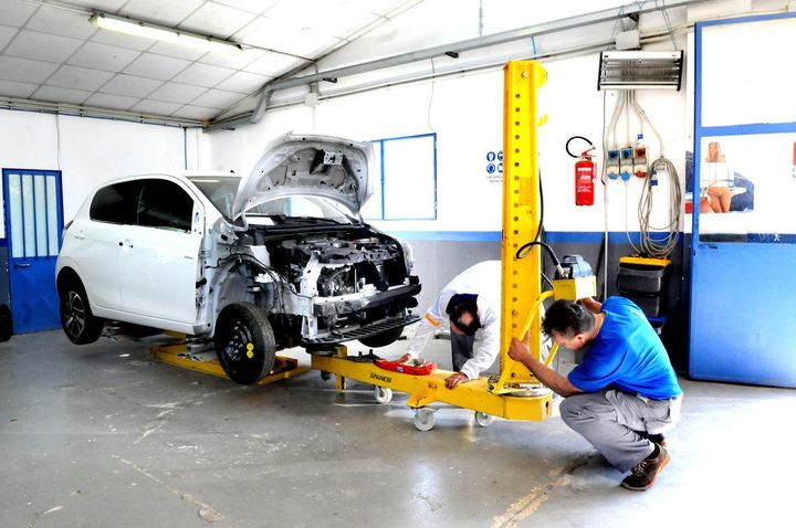Un SUV bianco viene riparato in un'officina, con un meccanico che utilizza un ponte sollevatore giallo vicino alla parte anteriore.