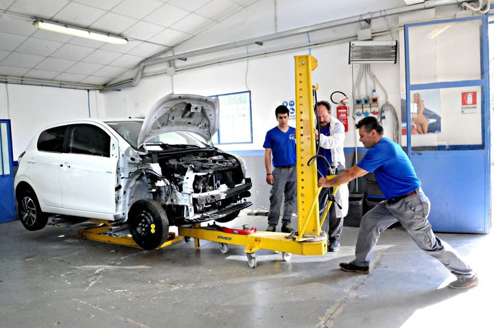 Un'auto bianca viene riparata in un'officina, con i meccanici che utilizzano un ponte sollevatore giallo e degli attrezzi.