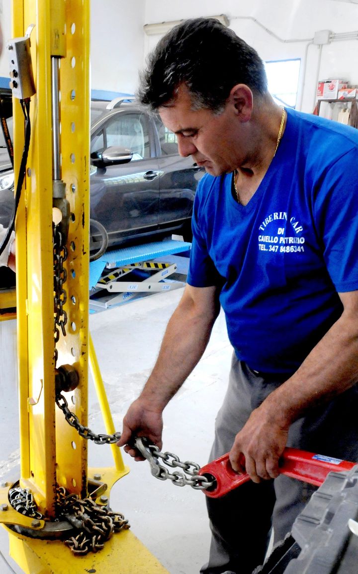 Tecnico con camicia blu che utilizza un attrezzo a catena su un ponte sollevatore giallo in un'officina.