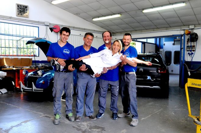 Cinque meccanici in uniforme blu trasportano un uomo sorridente con una camicia bianca all'interno di un'officina.