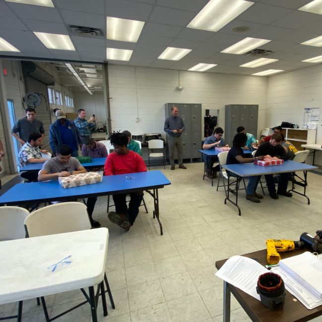 Classroom with students at blue tables, instructor standing. Students work on projects. Fluorescent lights illuminate room.