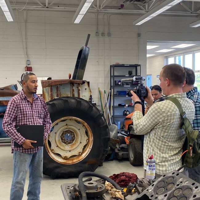 A man in a checkered shirt holds a laptop, standing near an old tractor being filmed in a workshop.