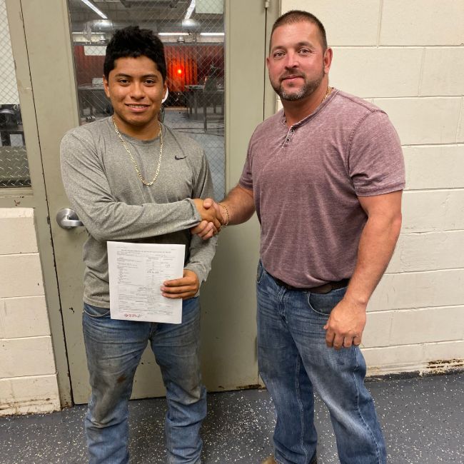 Two men shaking hands, one holding a paper. Indoor setting, gray and red tones.