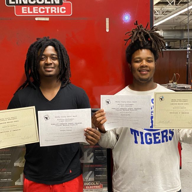 Two men holding welding certificates in front of a red background with Lincoln Electric branding.