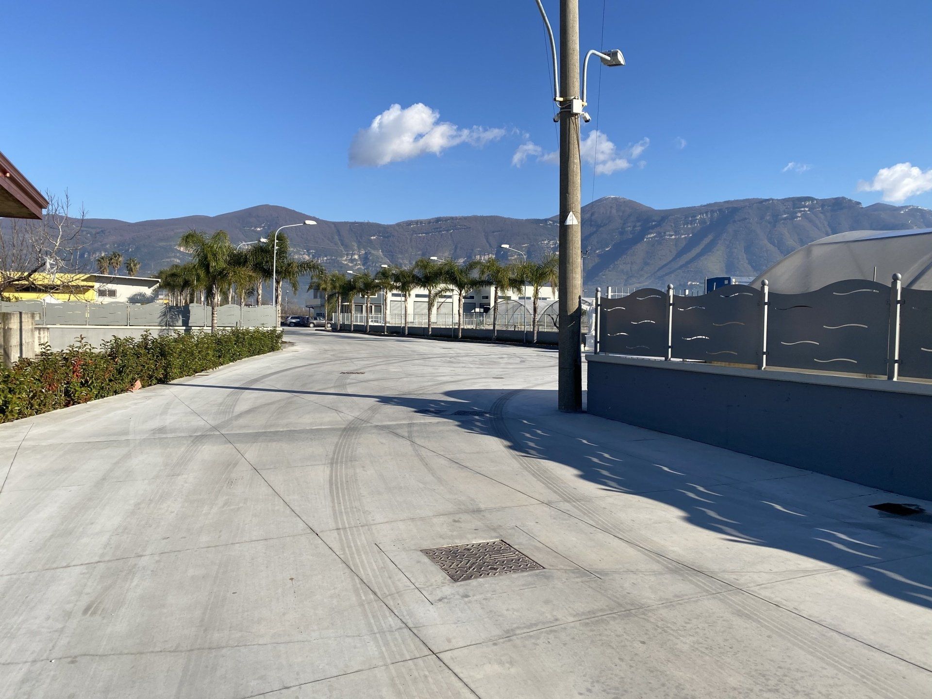 Strada asfaltata deserta accanto a una recinzione e a un palo della luce, con le montagne sullo sfondo sotto un cielo azzurro e limpido.