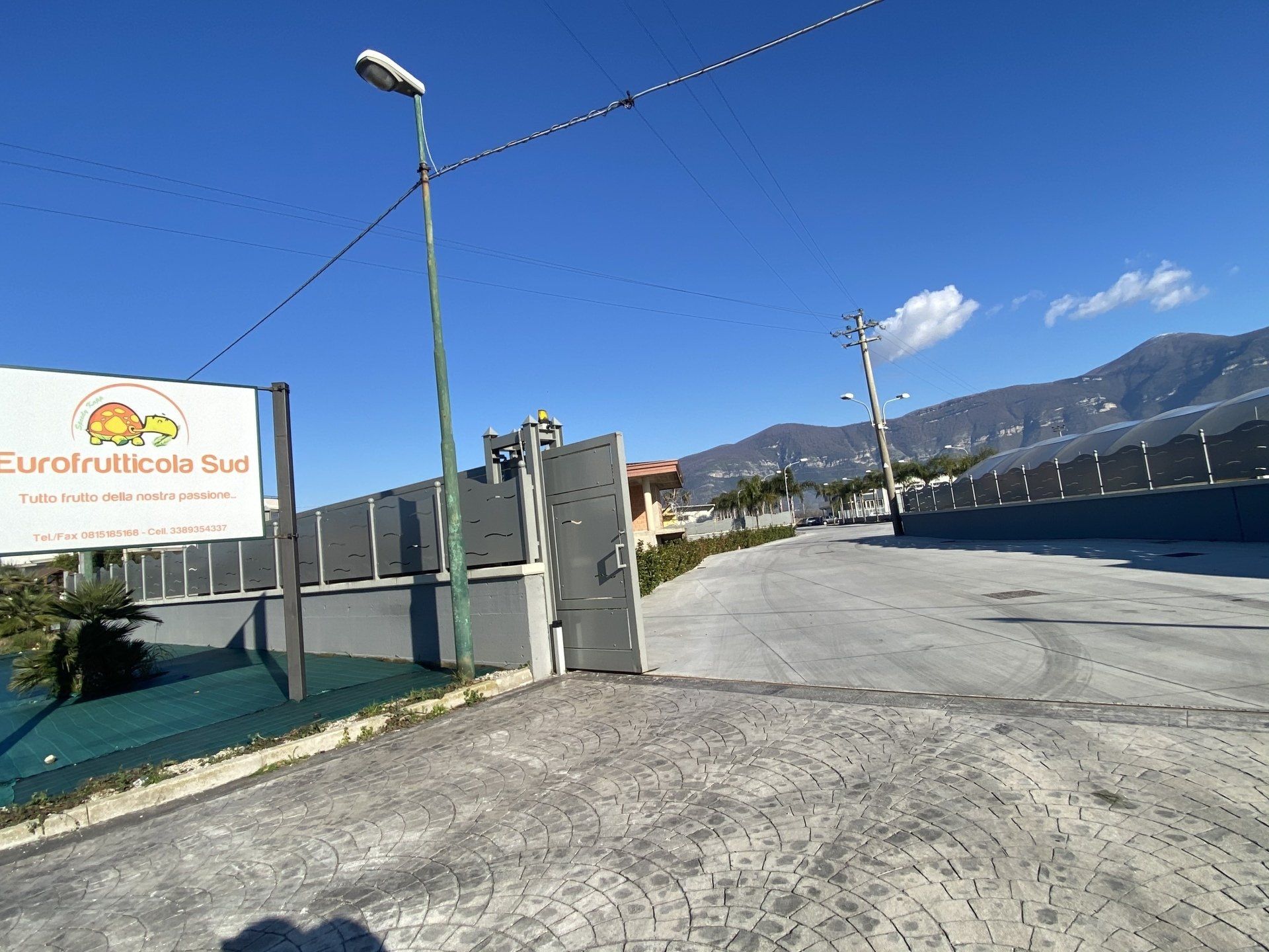 Ingresso carrabile con cancello e insegna, lampioni, montagne e un cielo azzurro in una giornata di sole.