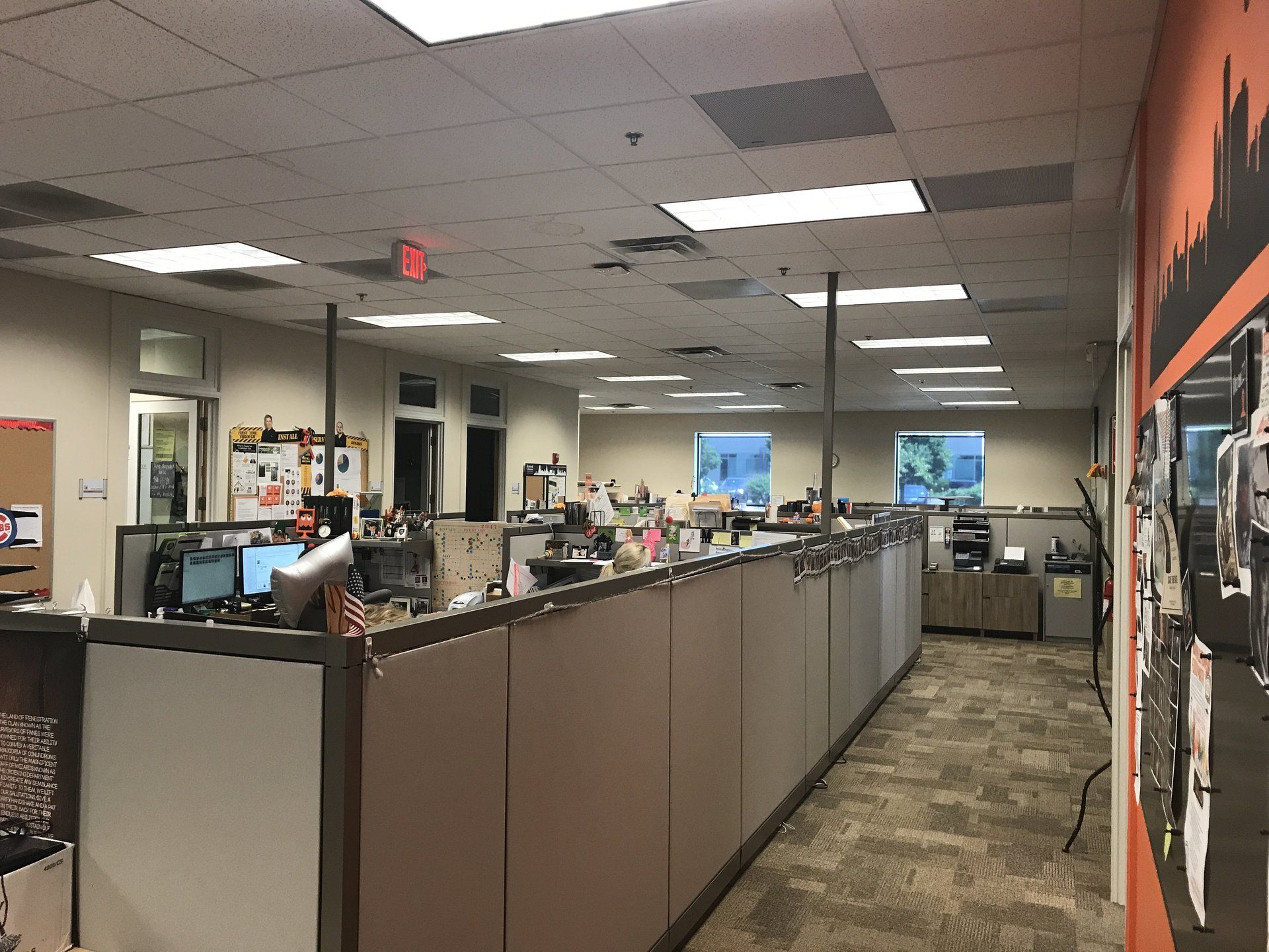 A large office with a lot of cubicles and a red exit sign on the ceiling.