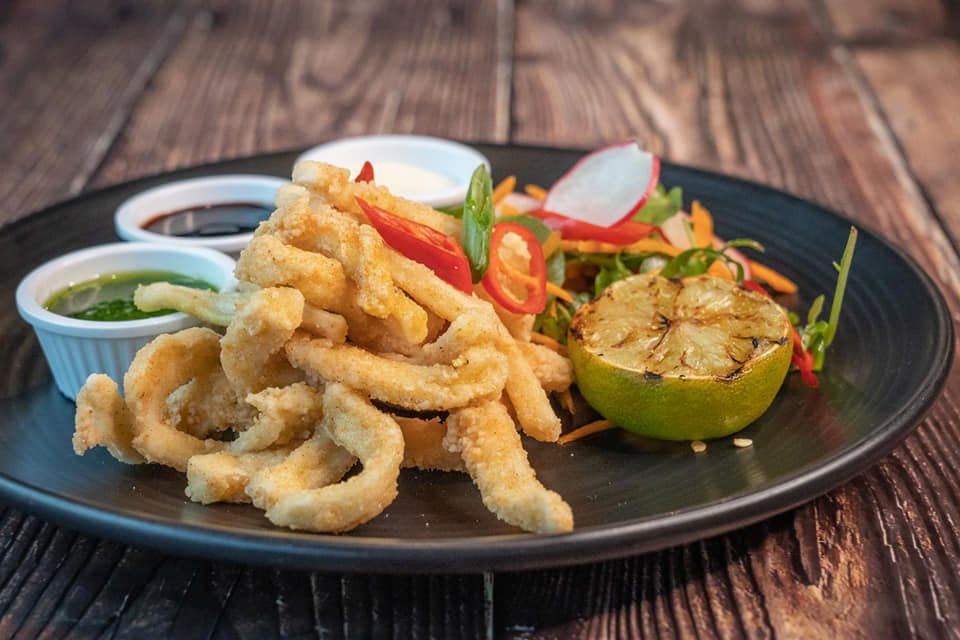 Fried calamari with dipping sauce and lime on a dark plate, garnished with herbs and sliced chilies.