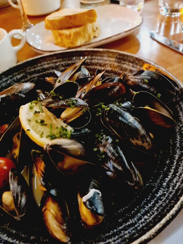 Plate of mussels in black broth with a lemon wedge on a restaurant table, with bread and drinks nearby