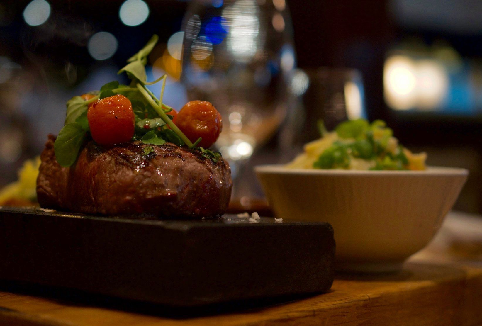 Gourmet steak topped with herbs and cherry tomatoes beside a side dish on a wooden table