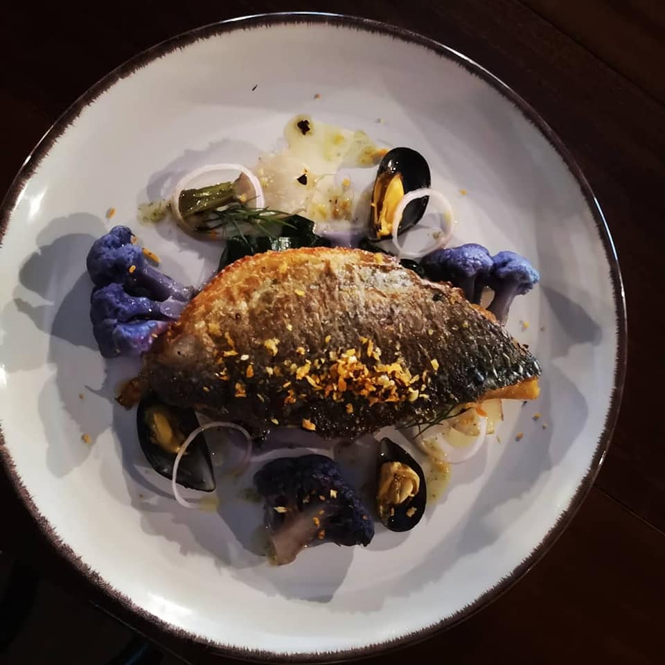 Seared fish on a white plate with mussels, purple garnish, and herbs