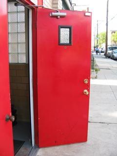 Red door, slightly ajar, with a small window. Outdoors, on a sidewalk near buildings and cars.