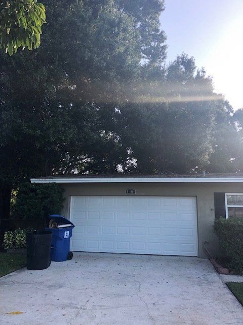 Garage with white door, trash cans, and large trees overhead; sun rays shine.