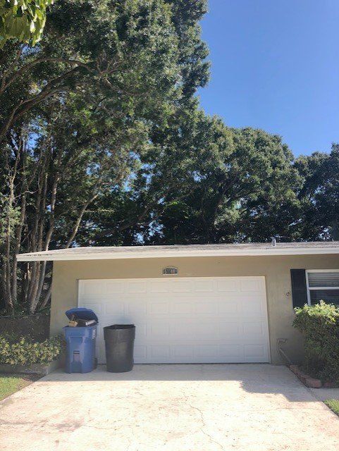 Tan garage with white door, blue and black trash bins, driveway, and lush green trees overhead.