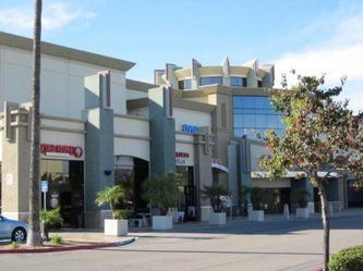 A shopping center with various storefronts, a blue sky, and a car in the parking lot.