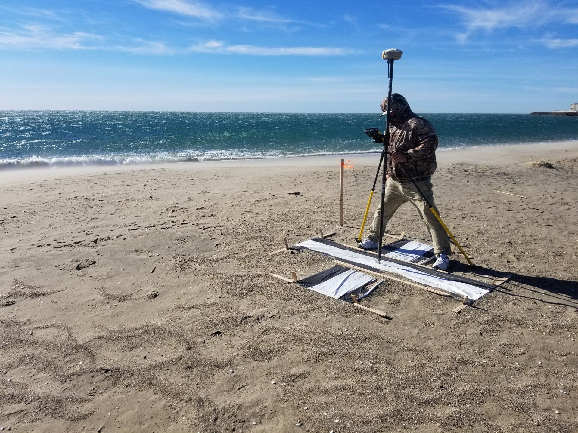Person using surveying equipment on a sandy beach near the ocean on a sunny day.