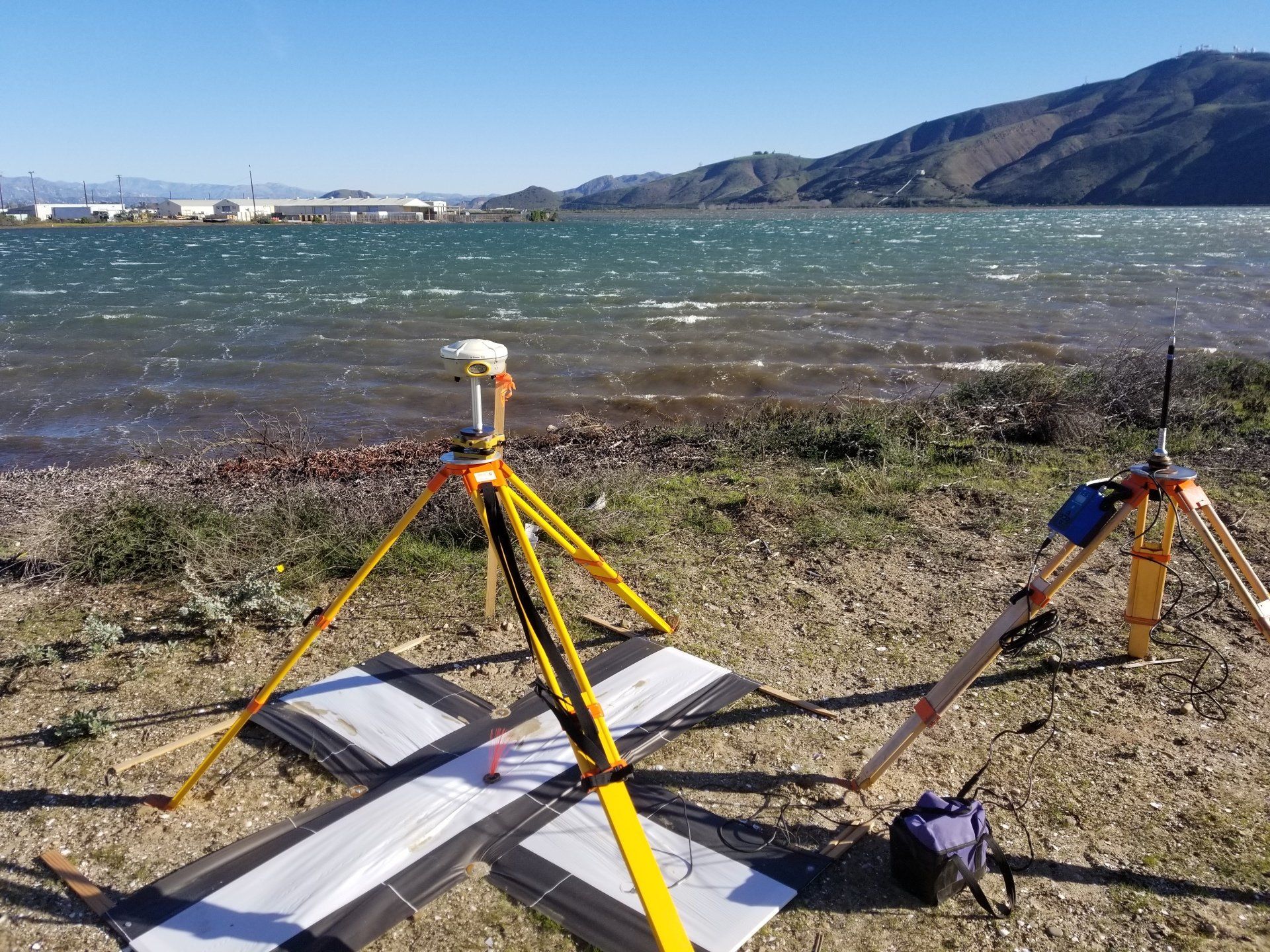 Surveying equipment by water, with a GPS receiver on a tripod, a black and white target, and mountains in the background.