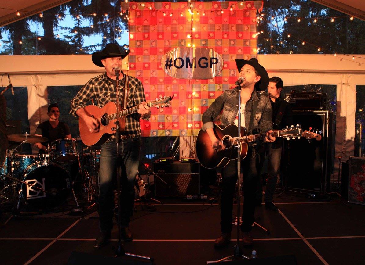 Two men are playing guitars on a stage in front of a sign that says #omgp