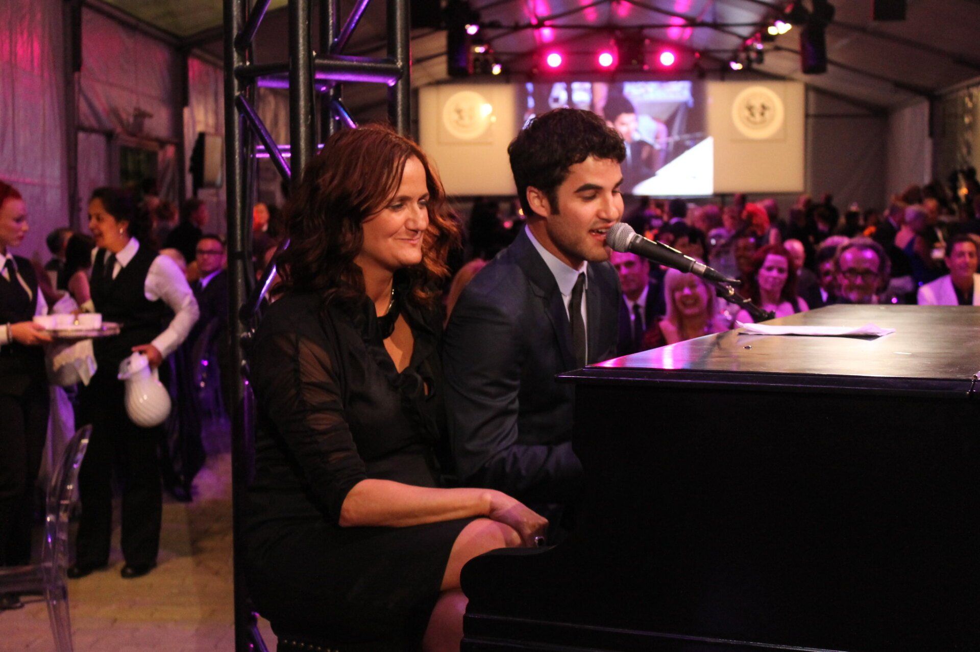 A man is singing into a microphone while a woman sits at a piano.