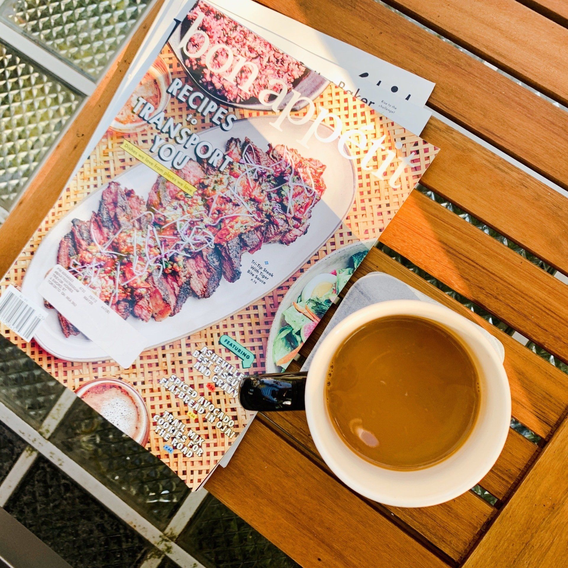A cup of coffee sits on a wooden table next to a stack of recipes magazines