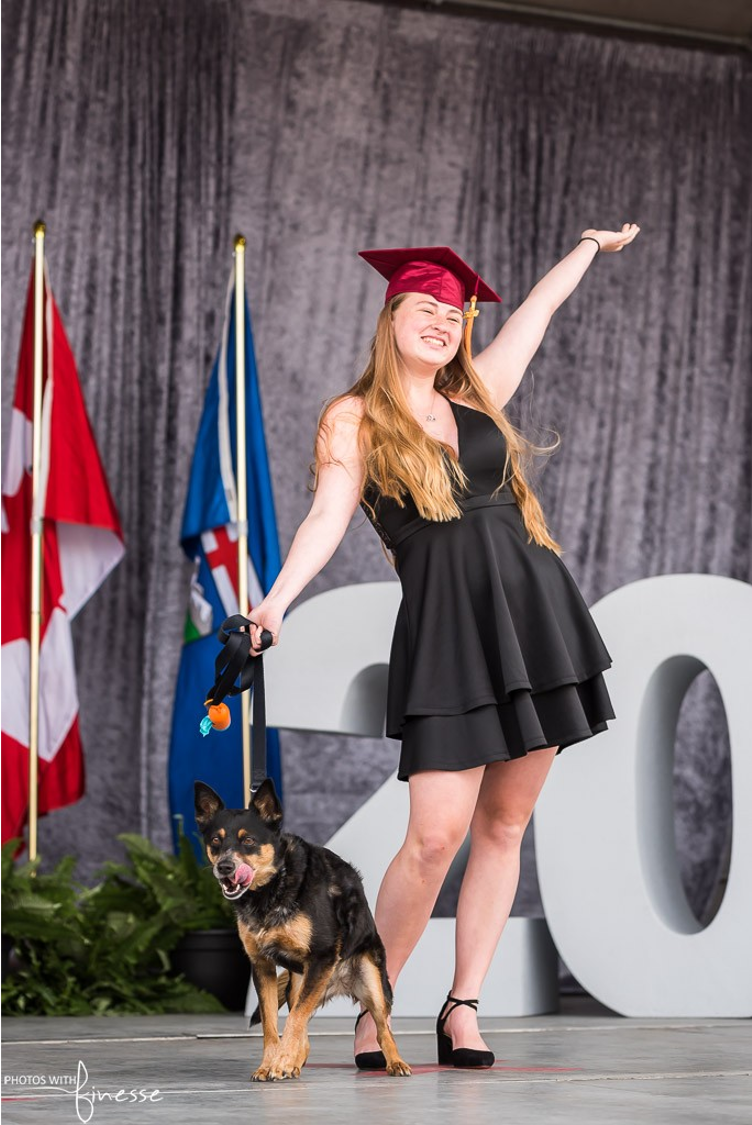 A woman in a graduation cap and gown is standing next to a dog.