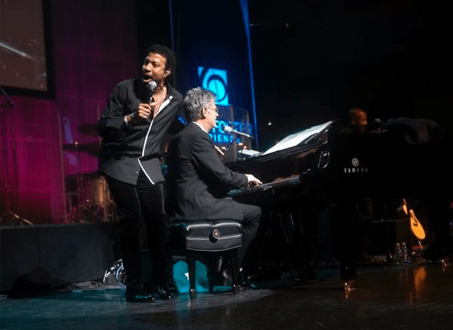 A man singing into a microphone next to a man playing a piano on a stage.