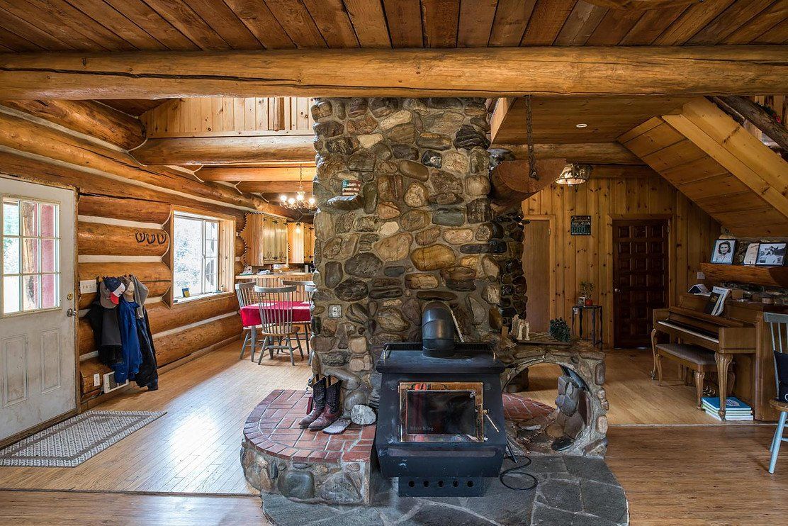 Rustic cabin interior with stone fireplace, wood stove, and log walls. View includes dining area, piano, and staircase.