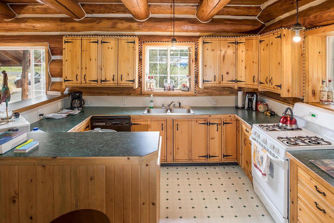 Log cabin kitchen with wood cabinets, green countertops, white stove, and window overlooking the outdoors.