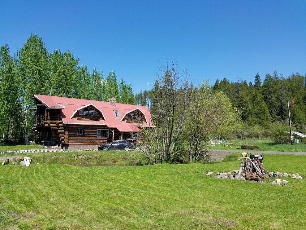 Log cabin with a red roof sits on a grassy lawn, trees in the background under a blue sky.