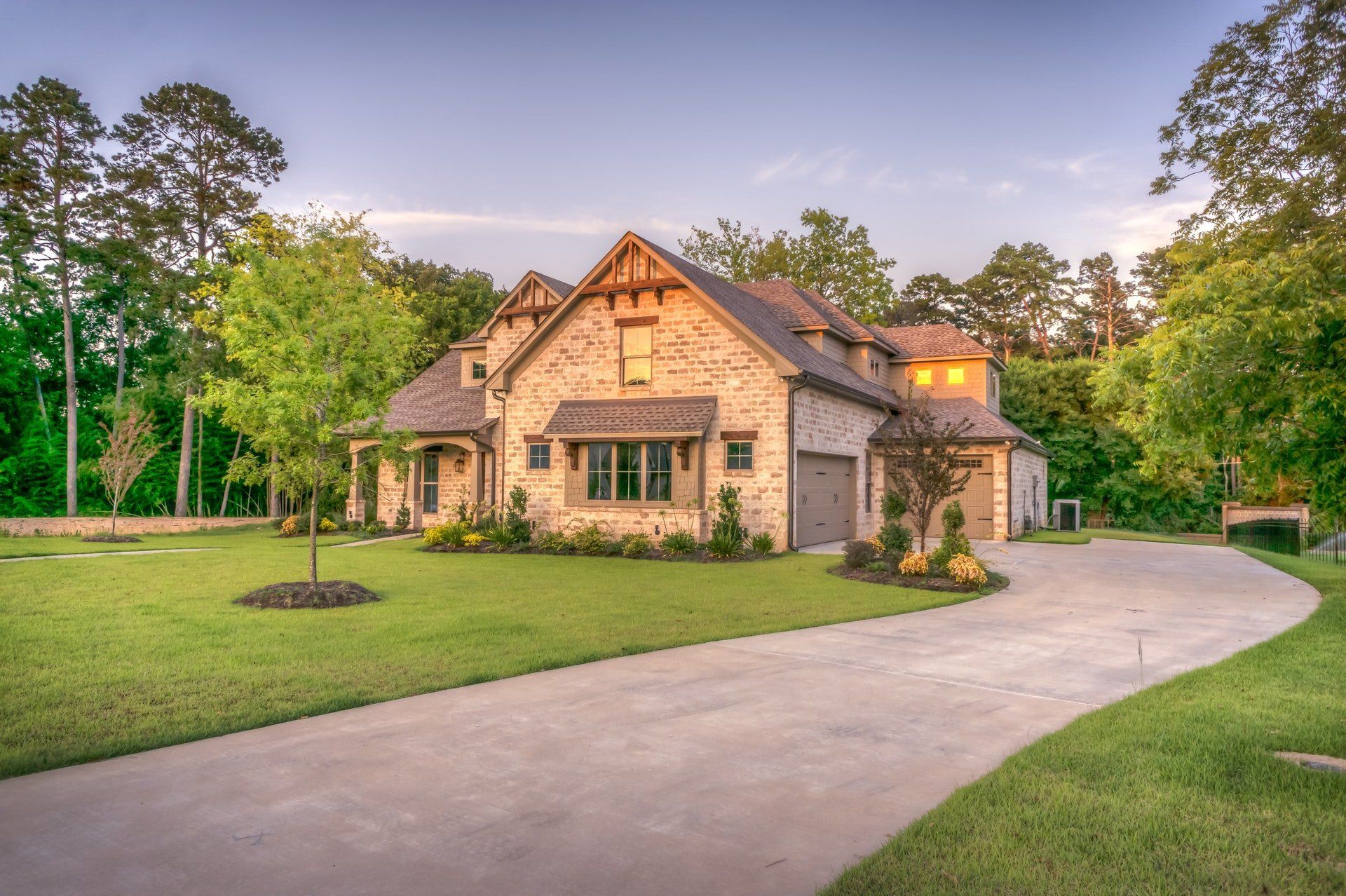 A stone-clad house with a winding driveway, surrounded by green lawn and trees, under a dusky sky.