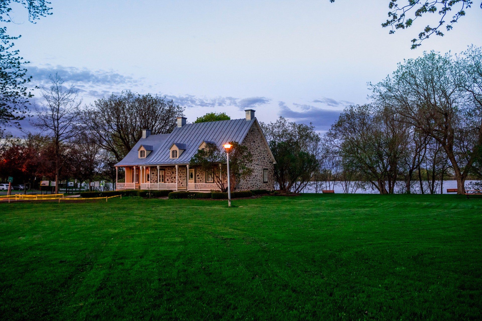 Stone house with a long porch and three dormers on a green lawn, trees, and a body of water under a dusk sky.