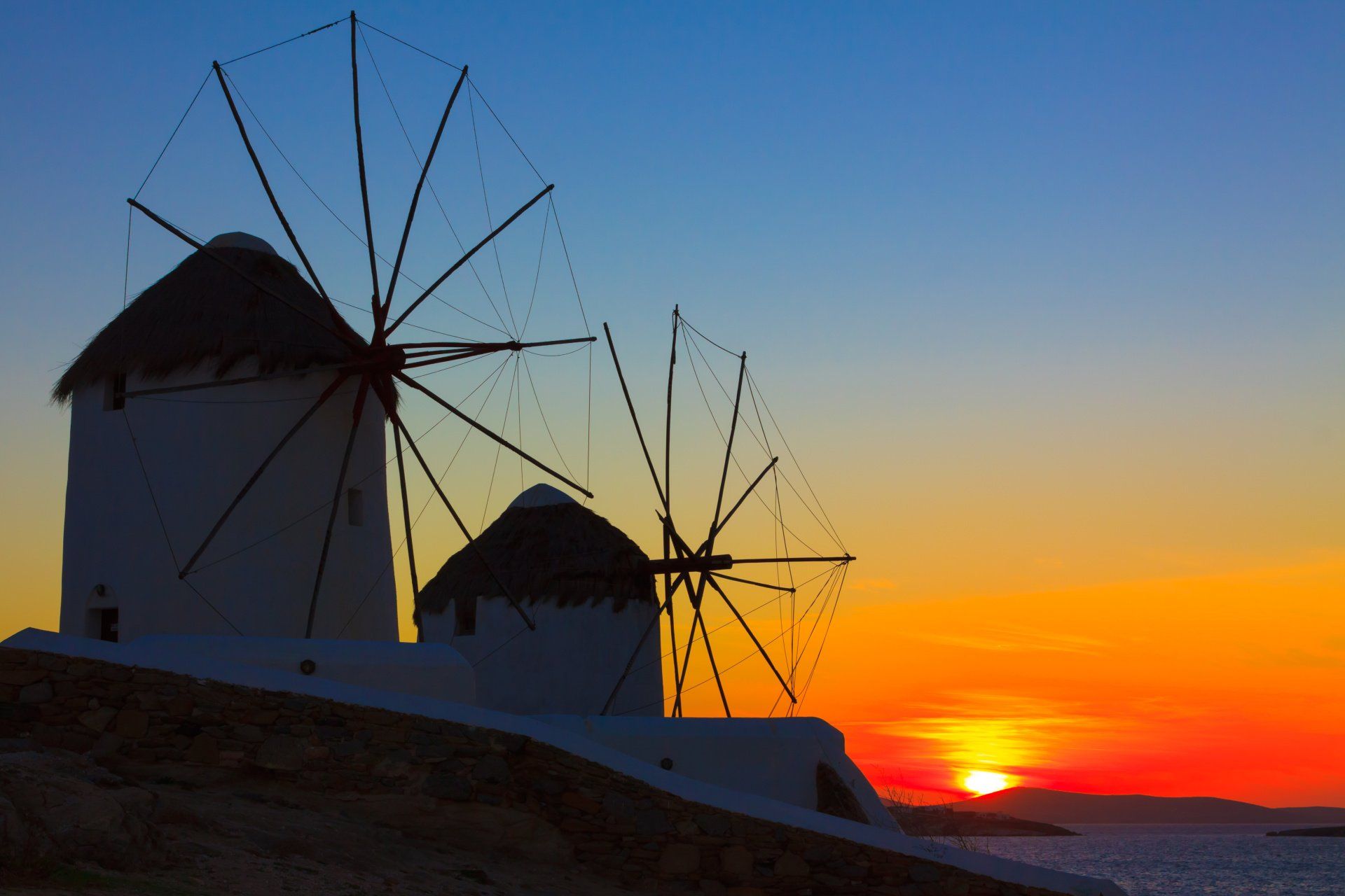 Two windmills are silhouetted against a sunset over the ocean