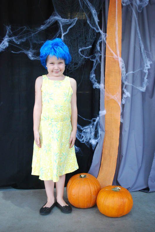 A little girl in a yellow dress and blue wig is standing next to pumpkins.