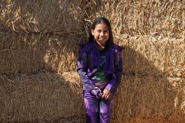 A little girl in a purple outfit is standing in front of a hay bale.