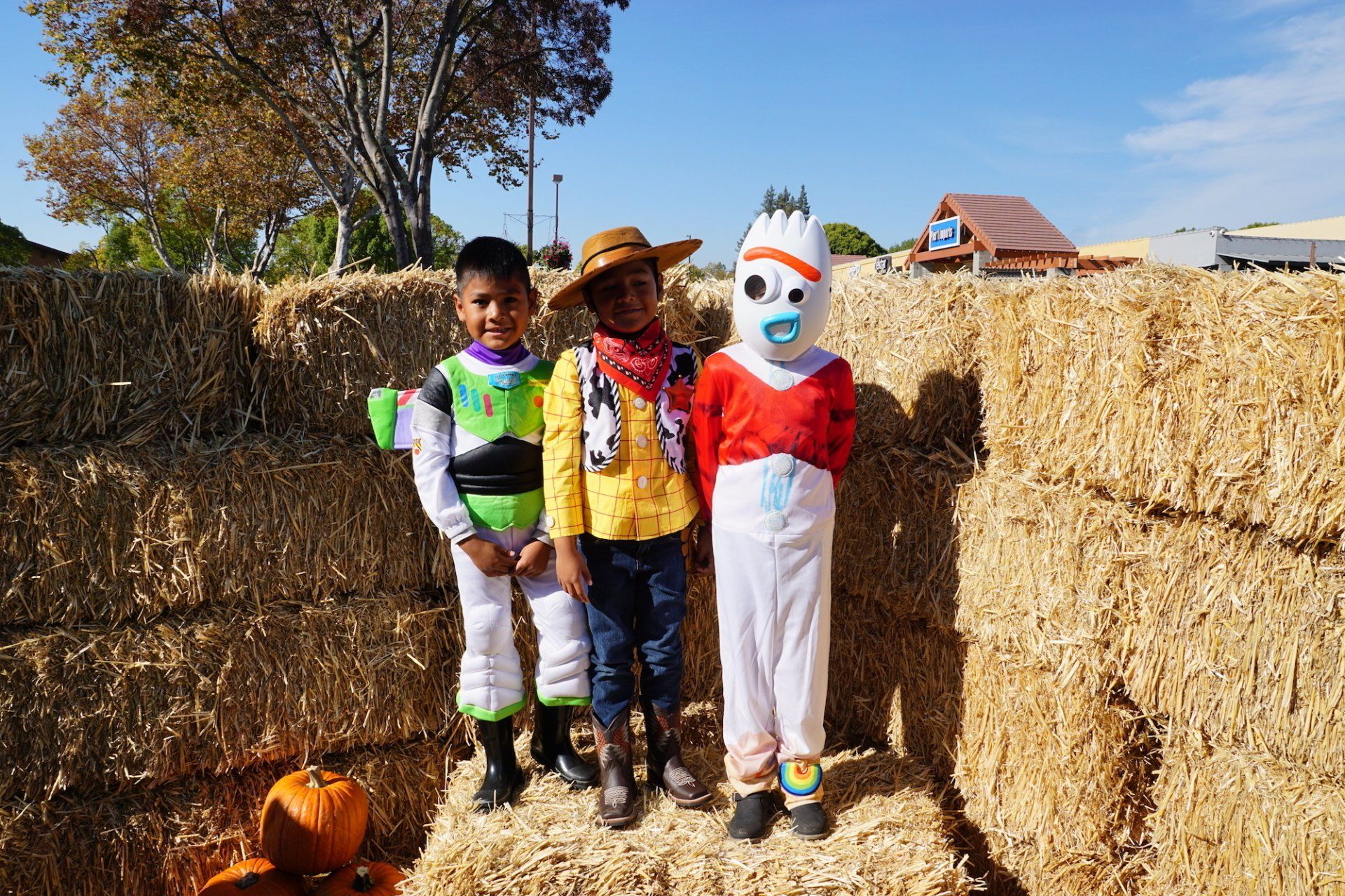Three children dressed in costumes are standing next to hay bales.