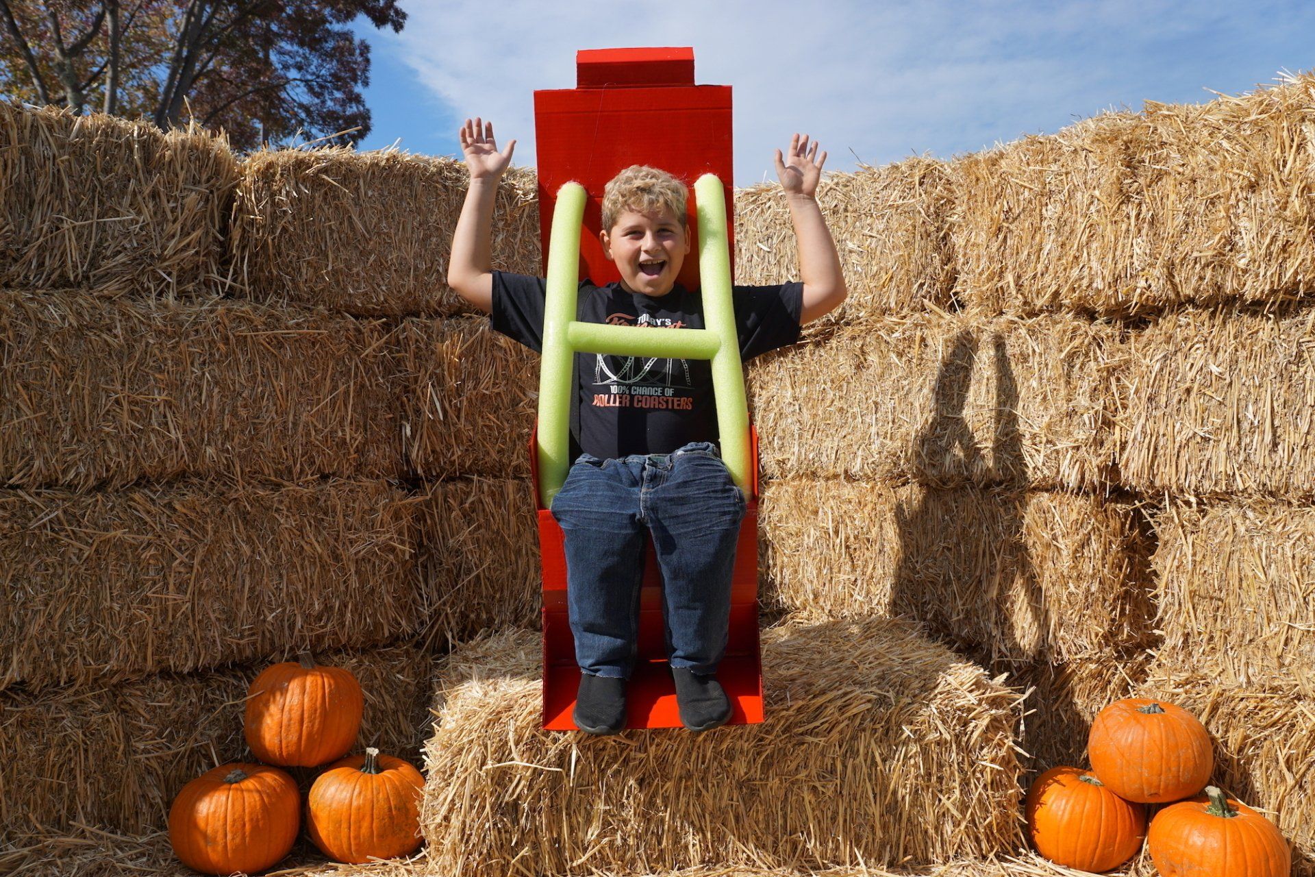 A young boy is sitting in a red chair surrounded by hay bales and pumpkins.