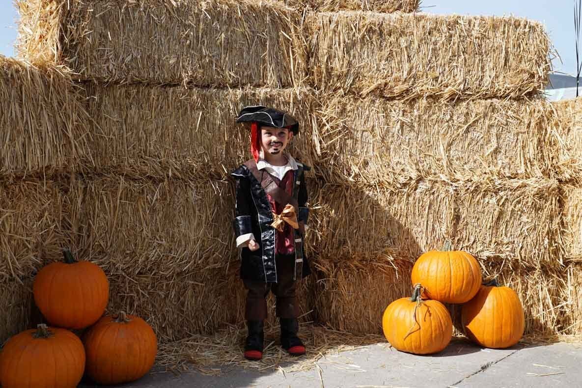 A little boy in a pirate costume is standing next to hay bales and pumpkins.
