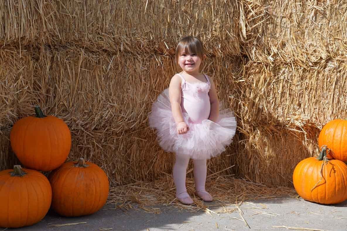 A little girl in a tutu is standing in front of a bale of hay surrounded by pumpkins.