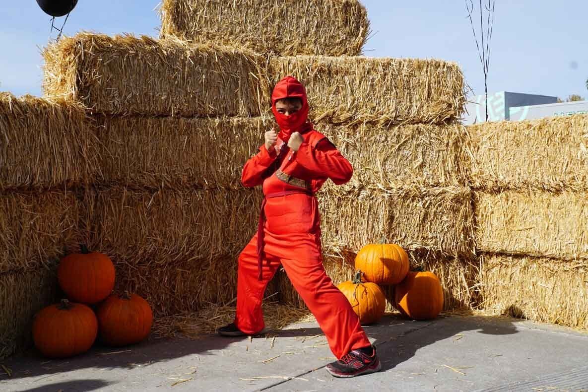 A man in a red ninja costume is standing in front of hay bales and pumpkins.