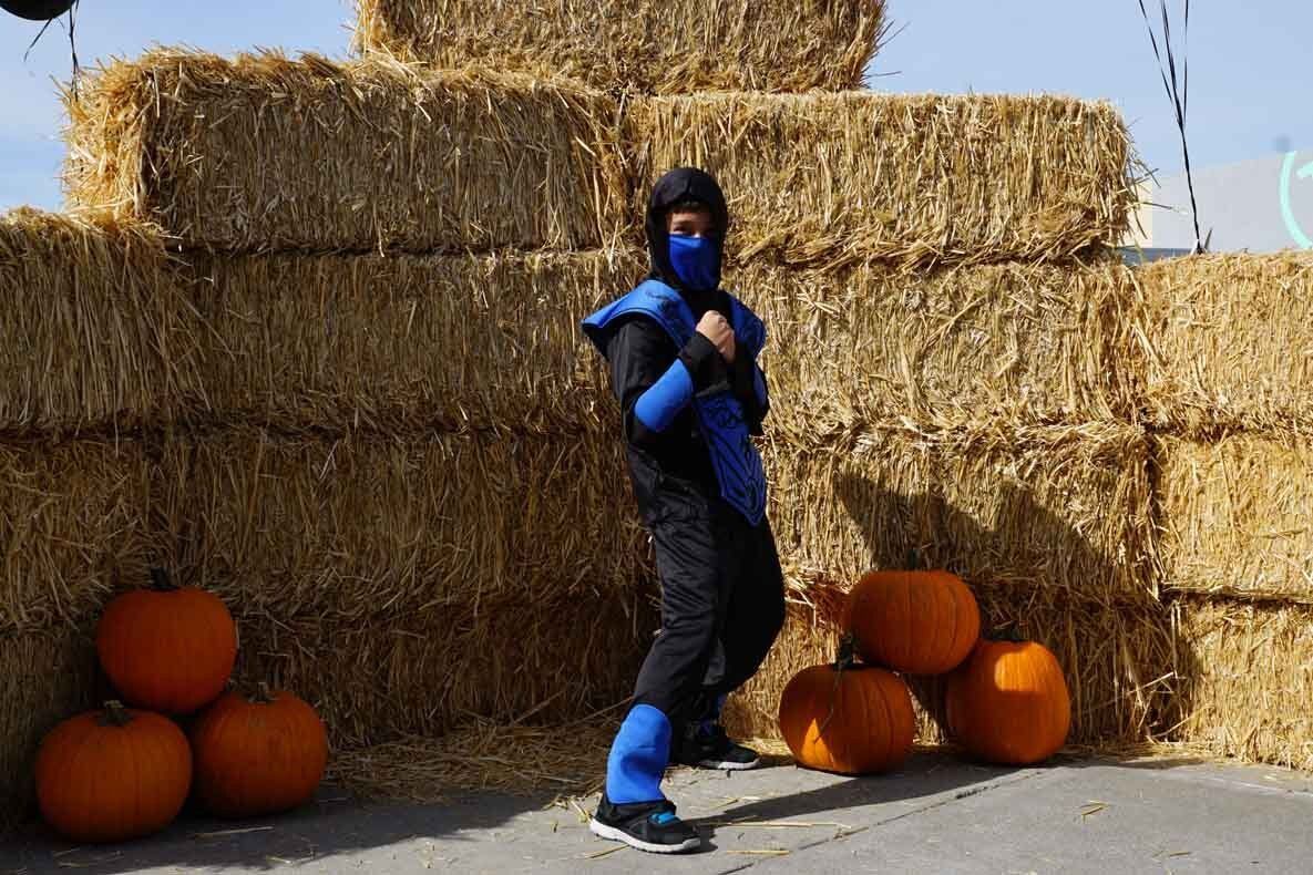 A person in a ninja costume is standing in front of hay bales and pumpkins.