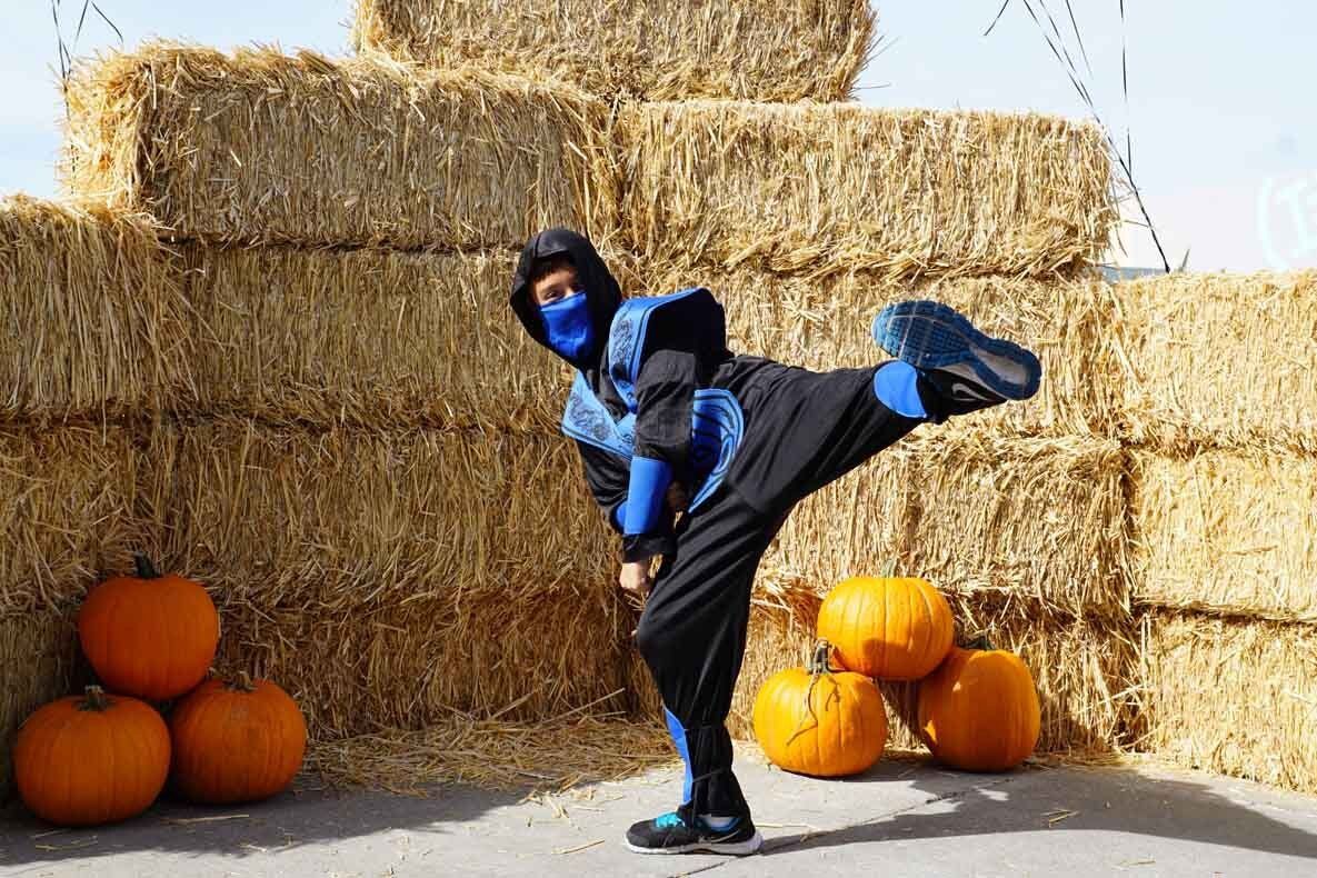 A person dressed as a ninja is kicking a pumpkin in front of hay bales.
