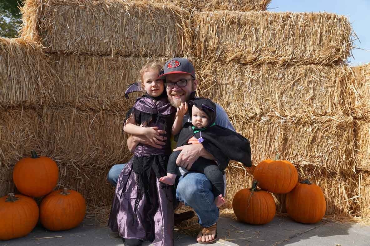 A man is holding two children in front of a pile of hay bales.