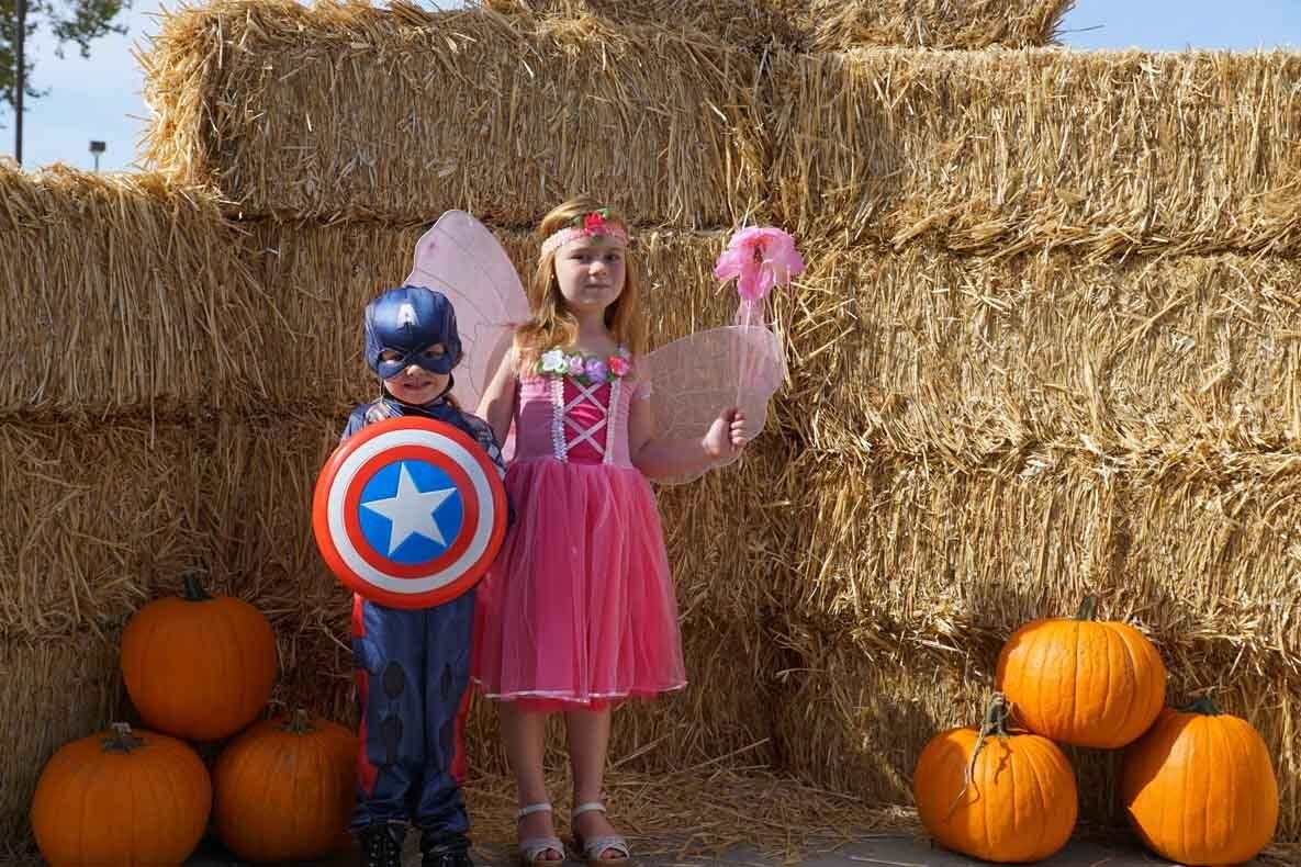 A boy in a captain america costume and a girl in a princess costume are standing next to pumpkins.