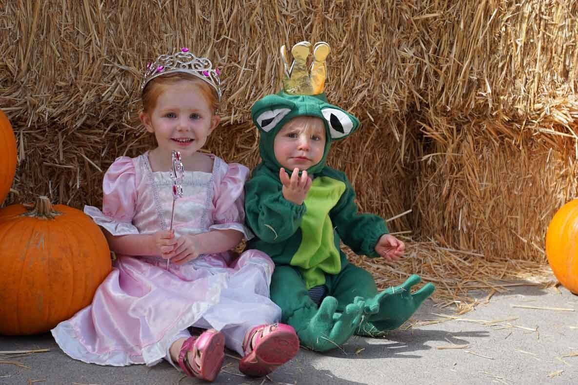 A little girl dressed as a princess and a little boy dressed as a frog are sitting next to pumpkins.